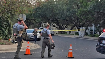 Dos agentes de policía con chalecos tácticos y cascos caminan cerca de conos de tránsito y cinta amarilla en una calle residencial