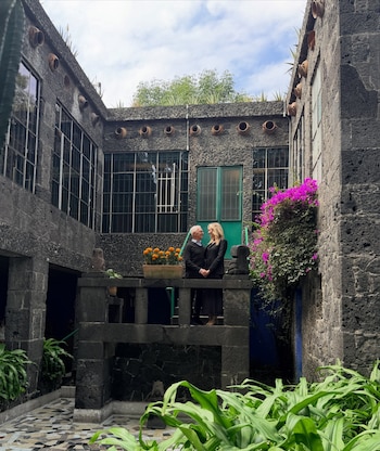 Un hombre y una mujer de pie tomados de las manos en un patio rodeado por muros de piedra volcánica, ventanas altas y plantas con flores