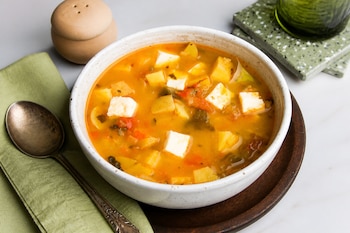 Un plato hondo blanco con caldo, cubos de queso, papas y chiles, acompañado de una cuchara sobre una servilleta verde.