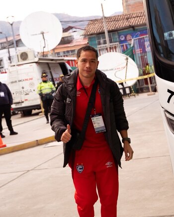 Hombre con chaqueta negra y camiseta roja con logo de Cienciano, pantalón rojo. Muestra el pulgar arriba. Lleva credencial de acceso. Fondo con bus blanco y camioneta con antenas