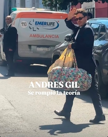 Un hombre con traje oscuro y gafas camina por la calle con bolsos y levanta una mano, delante de una ambulancia y un automóvil