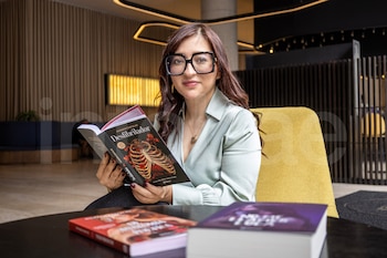 Una mujer sentada con lentes y blusa verde claro sostiene un libro titulado "Desfibrilador" de Gilraen Eärfalas; hay otros dos libros en una mesa