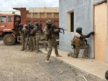 Varios militares con uniforme de camuflaje y cascos, uno arrodillado frente a una puerta de madera, y otros en fila con fusiles junto a un camión rojo