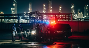 Cuatro policías en equipo táctico y un hombre con esposas junto a un vehículo SUV policial con luces rojas y azules intermitentes en un área industrial nocturna.