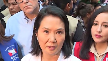 Mujer de cabello oscuro y blusa clara hablando frente a varias personas. A la izquierda, un hombre con gafas. A la derecha, una mujer con chaleco rojo