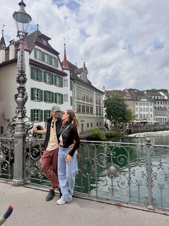 Pareja de pie en un puente de metal ornamentado, mirándose con cariño. Al fondo, edificios históricos con tejados rojos y un río de color verde turquesa