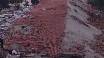 Techo de tejas rojas derrumbado, paredes de concreto rotas, gran cantidad de escombros de construcción y dos personas entre los restos de un edificio