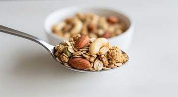 Cuchara de metal con granola, avena, almendras, anacardos y semillas de calabaza. Bol con granola desenfocado al fondo, sobre mesa clara.