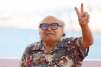 Danny DeVito con gafas de marco negro y camisa estampada levanta los dedos en señal de victoria