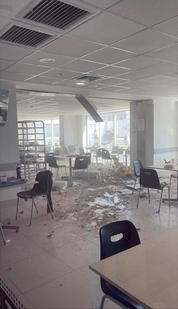 Interior de un gran espacio con baldosas claras, cubierto de escombros grises y blancos de techo colapsado. Sillas negras y mesas están dispersas, algunas con desechos