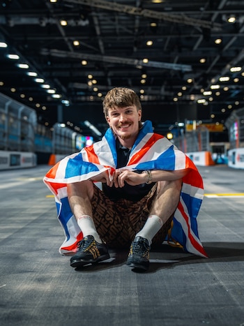 Un hombre sonriente sentado en el suelo de una pista cubierta, envuelto con la bandera del Reino Unido