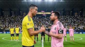 Cristiano Ronaldo con camiseta amarilla y Lionel Messi con camiseta rosa se estrechan la mano de perfil en un estadio iluminado.