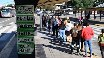 Un cartel de Metrobús de la línea 45 con destinos como Obelisco y Retiro en primer plano, con personas esperando en la parada y un colectivo en la calle de fondo
