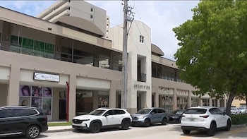 Vista frontal de edificios comerciales en Hialeah, con tiendas a pie de calle, varios autos estacionados y un árbol frondoso a la derecha. Un poste de servicios se alza en el centro