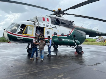 Tres agentes policiales escoltan a un hombre esposado frente a un helicóptero blanco y verde de la policía en una pista mojada