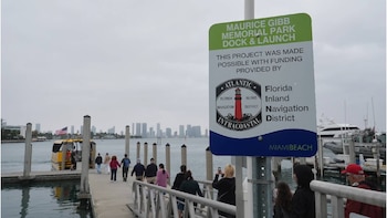 Una señal de parque y un muelle con personas caminando hacia un barco amarillo con la bandera de EE. UU. en Miami Beach; un horizonte urbano al fondo