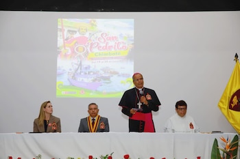 Un obispo habla en un podio con micrófono. Dos personas están sentadas a una mesa frente a él. Detrás, una pantalla proyecta el cartel de la 'Festividad de San Pedrito Chimbote'