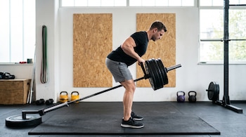Hombre caucásico realiza remo T-bar con barra y discos. El gimnasio tiene suelo de goma, ventanas, mancuernas, pesas rusas y bandas de resistencia.