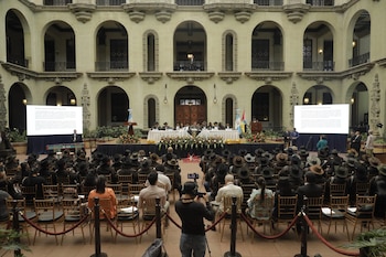 Amplio salón interior con arcos y balcones. En el centro, un escenario con mesa, dos pantallas, banderas y personas. Audiencia en sillas de madera