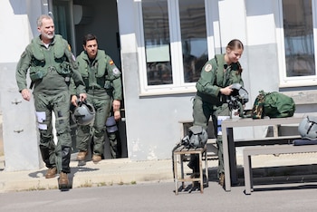 Rey Felipe VI, Princesa Leonor y un hombre con trajes de vuelo verdes. Salen de una puerta y ajustan cascos de aviación junto a una mesa y un taburete
