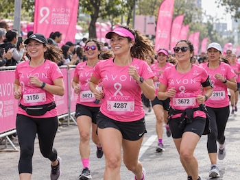 Varias mujeres con camisetas rosadas y dorsales numerados corren por la calle junto a banderolas rosas