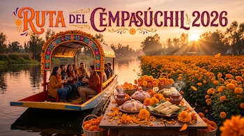 Personas en una trajinera navegan un canal junto a una mesa con pan de muerto, tamales, bebidas y flores de cempasúchil al atardecer.