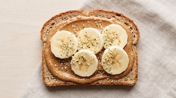 Una rebanada de pan tostado con mantequilla de almendra, cinco rodajas de banana y semillas de cáñamo sobre una superficie de madera clara y una servilleta beige.