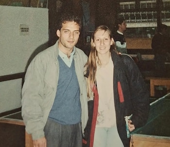 Un hombre joven con chaqueta gris y suéter azul junto a una mujer de abrigo oscuro en un recinto interior