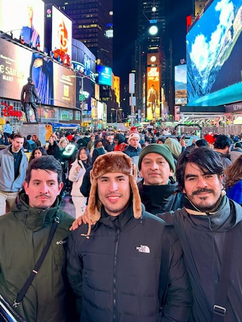 Cuatro hombres posan sonriendo en Times Square, Nueva York, de noche. Detrás de ellos, una multitud y grandes pantallas luminosas con publicidad