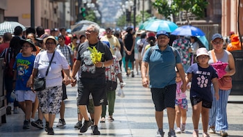 Varias personas caminan por una calle peatonal soleada usando gorras, sombreros y sombrillas para protegerse del sol