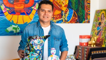 Un hombre con camisa de mezclilla sostiene una escultura pintada de un perro sin pelo, frente a cuadros coloridos y figuras sobre una mesa