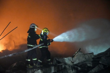 Dos bomberos con cascos y mascarillas apuntan una manguera con un potente chorro de agua hacia un gran incendio que emite un resplandor naranja en la noche