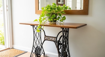Mesa hecha con base de hierro forjado de máquina de coser Singer antigua, con planta verde en maceta y espejo de madera. Pared blanca, felpudo.