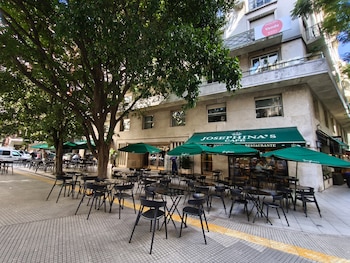 Fachada de Josephina's Café con toldos y sombrillas verdes, múltiples mesas y sillas negras en la acera, árboles frondosos y un edificio con cartel de venta