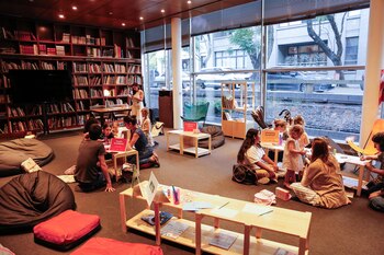 Estanterías con libros, pantalla de televisión, mesas bajas de madera, cojines y pufs, adultos y niños sentados, grandes ventanales a la calle, letreros