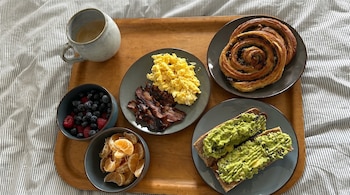 Bandeja de madera con taza de café, huevos revueltos, tocino, tostadas de aguacate, bollo, arándanos, frambuesas y gajos de mandarina.