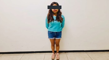 Mujer de pie con suéter azul, pantalón corto de mezclilla, zapatillas rosadas y una barra negra sobre los ojos ante una pared blanca.