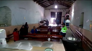 Interior de iglesia rural con paredes blancas y techo de madera. Varias personas, una con chaleco reflectante, observan bancas de madera y un altar