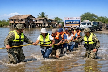 Dos militares y siete civiles con chalecos salvavidas cruzan agua con una cuerda. Detrás, casas elevadas, vehículos y un cartel de simulacro de inundación.