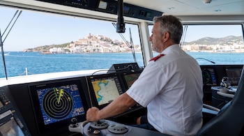 Un capitán con uniforme opera controles en el puente de mando de un yate, con pantallas de radar y cartas náuticas. La costa de Ibiza es visible al fondo.