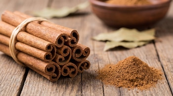 Manojo de ramas de canela Cassia atadas con cuerda sobre una mesa de madera, junto a un montículo de canela en polvo y hojas secas.