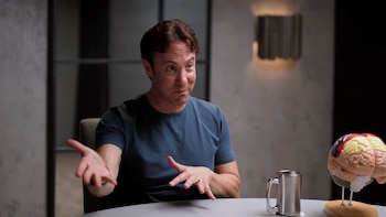 El Dr. David Eagleman, un hombre de cabello castaño con una camiseta azul, gesticula mientras está sentado en una mesa con un modelo cerebral y una taza de metal