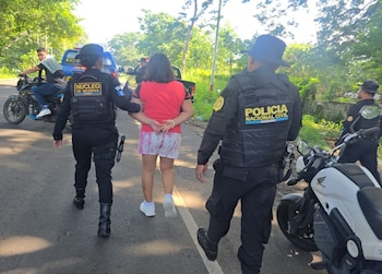 Dos agentes de policía guatemalteca escoltan a una mujer esposada en una carretera; se observan motocicletas y un vehículo pick-up en un entorno rural