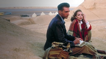 Ralph Fiennes y Kristin Scott Thomas sentados en el desierto. Él está de perfil, escribiendo en un libro. Ella lo mira con un chal rojo