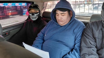 Tres hombres sentados en el asiento trasero de un coche. El hombre del centro lleva una sudadera azul y sostiene papeles. El hombre de la izquierda viste una máscara de calavera