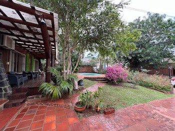 Jardín de una propiedad con galería de madera, patio de baldosas rojas, césped, plantas con flores rosadas y una pequeña piscina al fondo