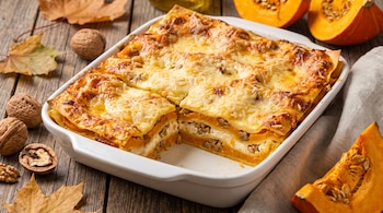 Una fuente blanca de lasaña de calabaza, ricota y nueces, con queso gratinado, sobre madera con hojas secas, nueces y trozos de calabaza.