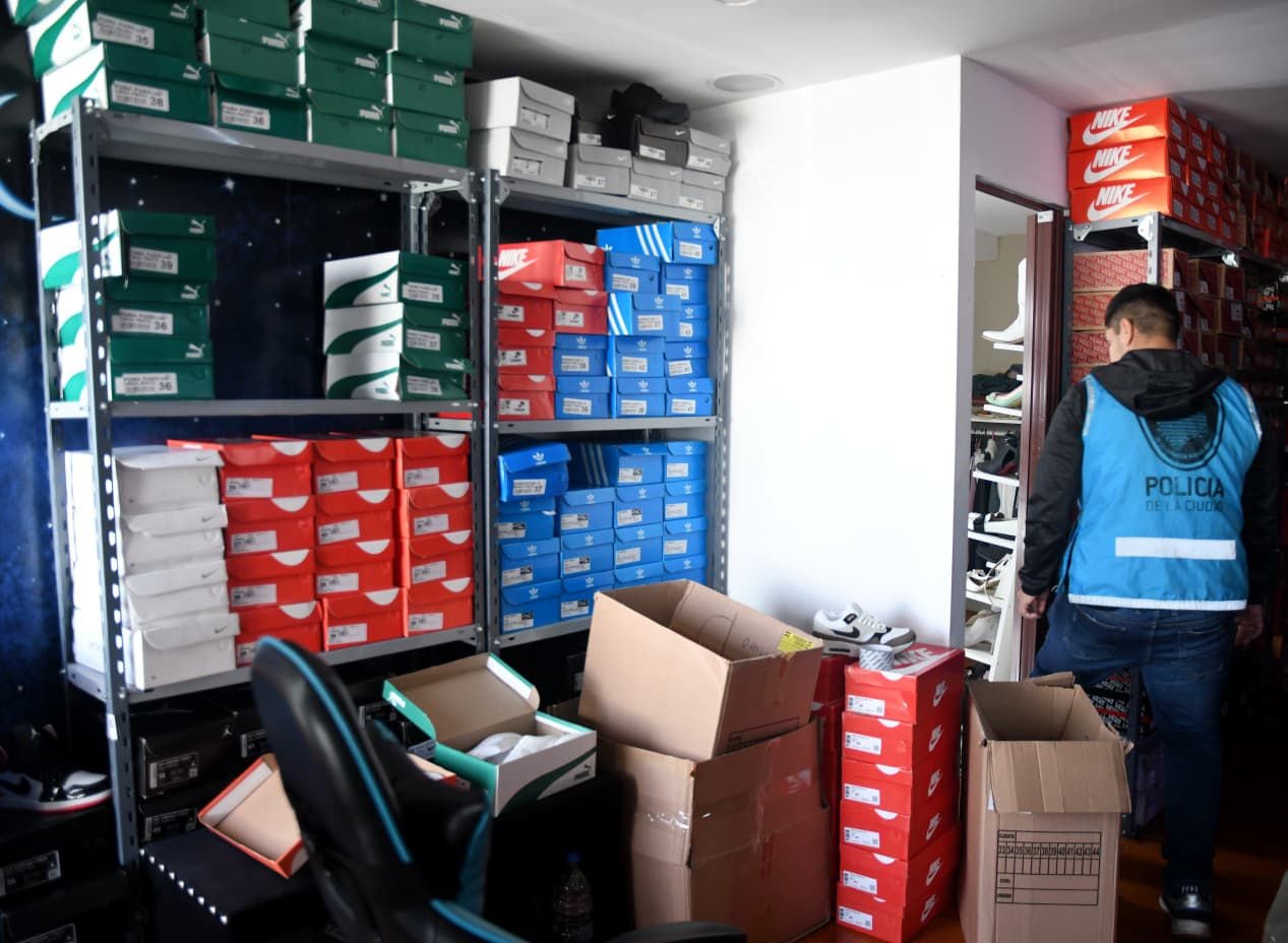 Un oficial de la Policía de la Ciudad inspecciona un depósito repleto de cajas de zapatillas de marcas como Nike y Adidas, incautadas en un operativo contra la falsificación.