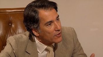 Primer plano de un hombre maduro con traje beis, camisa blanca y corbata, junto a un sillón de cuero marrón.