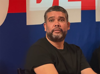 Primer plano de un hombre de mediana edad con cabello y barba grises, vistiendo una camiseta negra, con expresión seria y la boca ligeramente abierta, sobre un fondo bicolor rojo y azul